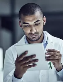 Male scientist in white lab coat, sitting next to microscope, looking at tablet.