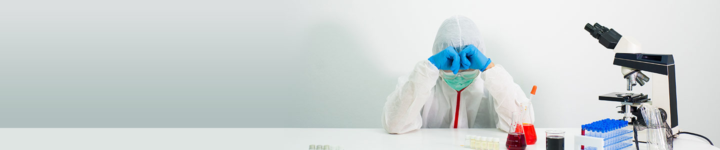 Scientist wearing PPE, sitting with elbows on the table covering his eyes as if disappointed with a microscope, test tubes, and beakers nearby.