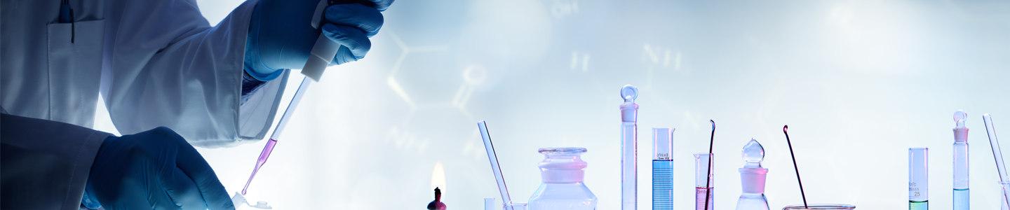 Scientist using pipette to put pink liquid into small tube, bottles full of various colored liquids on table nearby.