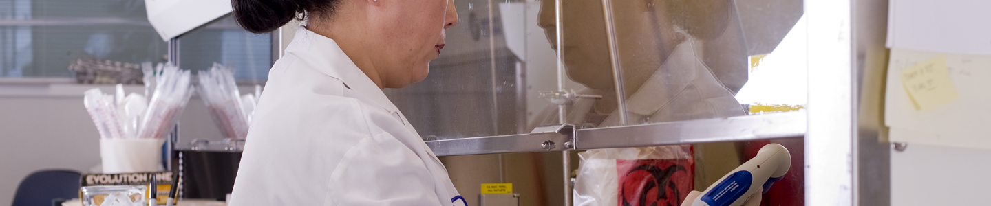 Female ATCC scientist in lab coat, sitting at biosafety cabinet, with hands under hood, pipette controller in hand.