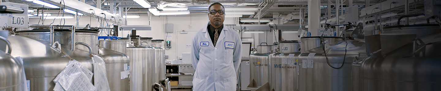 Scientist wearing lab coat, facing camera, standing between rows of large cryopreservation storage tanks at biorepository.