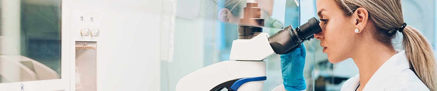 Female scientist in lab coat and gloves at workstation in laboratory, looking in microscope.