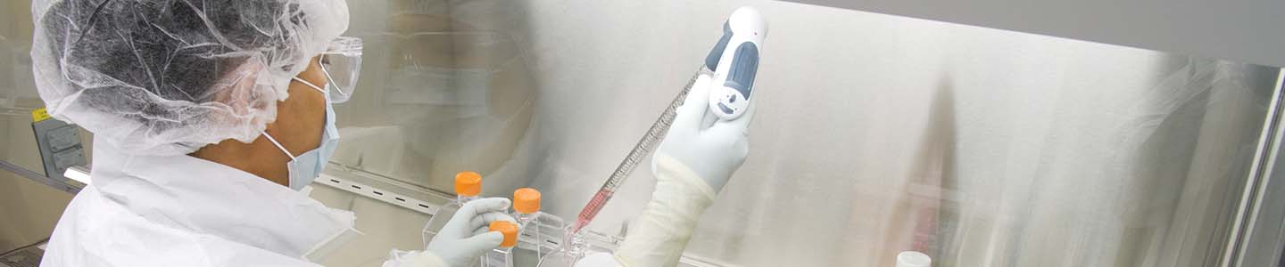 Side view of ATCC scientist wearing hair net, mask, smock, safety glasses, and gloves, holding a pipette aid with stopper and media bottle under a biological safety cabinet hood in a lab.