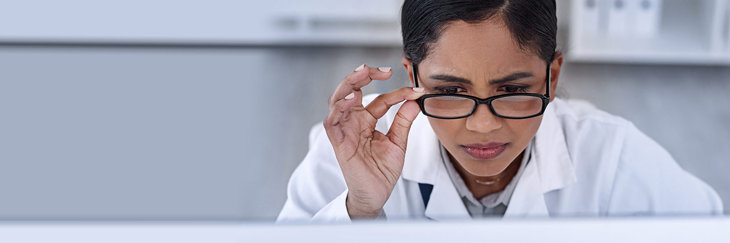 Female scientist looking down at a computer screen, holding one side of her eye glasses, as if confused.