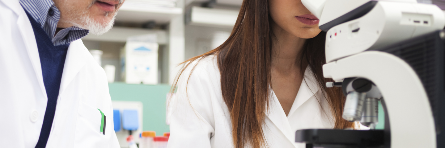 Scientist holding pen standing next to scientist looking into microscope.