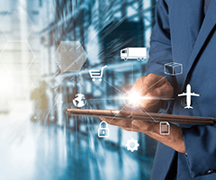 Man in a suit standing in a warehouse holding an iPad with light and symbols like an airplane, truck, globe, and shopping cart floating above his finger.