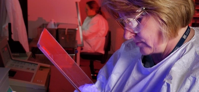 A person in a lab coat and gloves uses a pipette to transfer liquid into a multi-well plate under blue light. Another lab worker is visible in the background, also in lab attire.