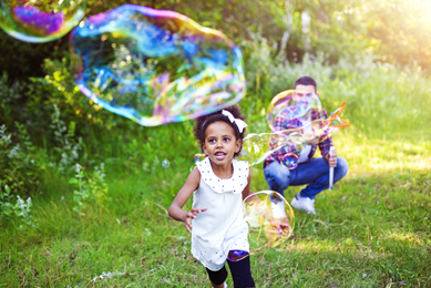 A young Black girl with a ribbon in her hair running outside in the grass toward a huge elongated bubble while a smiling man with a bubble blower squats in the background.