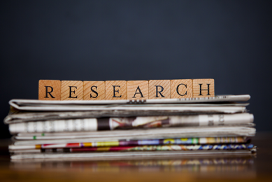 A row of small brown blocks, each with a letter, that spells "RESEARCH" on top of a stack of newspapers that appear blurry.