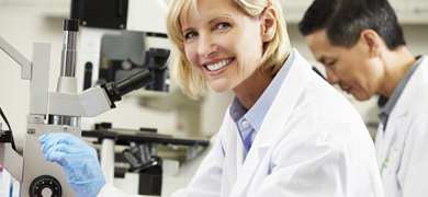 Female scientist wearing lab coat and gloves smiling at camera, at microscope holding a slide.