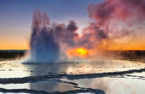 A plume of smoke rising from layers of slate-like liquid at Great Fountain Geyser in Yellowstone National Park at sunset.