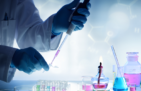 Scientist using pipette to put pink liquid into small tube, bottles full of various colored liquids on table nearby.