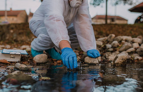 Person in personal protective equipment squatting and reaching to collect a water sample from a stream.