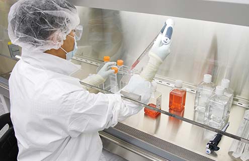 Side view of ATCC scientist wearing hair net, mask, smock, safety glasses, and gloves, holding a pipette aid with stopper and media bottle under a biological safety cabinet hood in a lab.