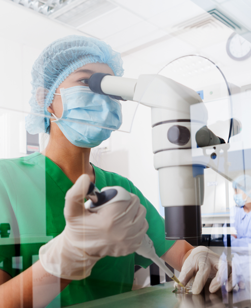 Scientist wearing surgical smock, cap, mask, and gloves, looking into microscope while using pipette.