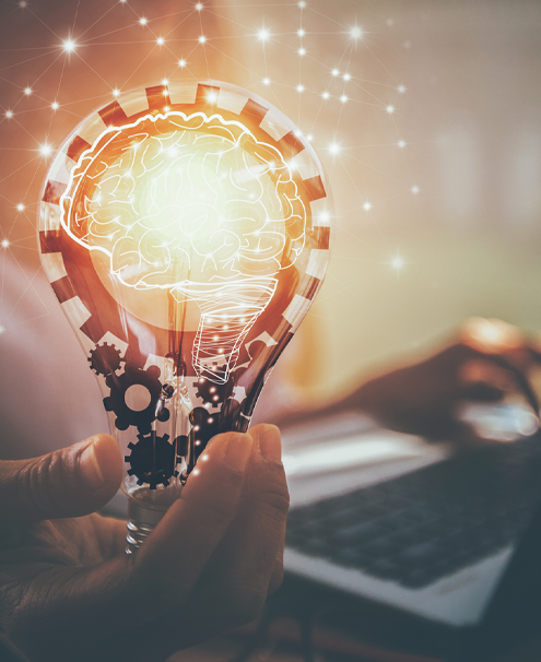 A person sitting in the background with one hand on a computer keyboard and the other hand holding an illustration of a light bulb, with gears, filament and white outline of a human brain with sparks of light connected by lines around it.