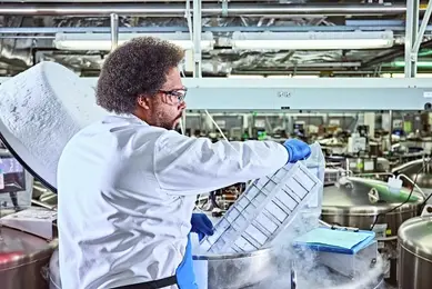 A laboratory technician in a white coat and blue gloves handles a tray of samples above a large stainless steel cryopreservation tank emitting vapor, likely from liquid nitrogen, with repository equipment visible in the background.