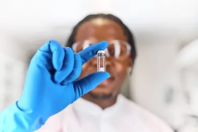Out of focus black, male, scientist wearing a blue glove holding a Microquant vial infront of their face that is in focus. 