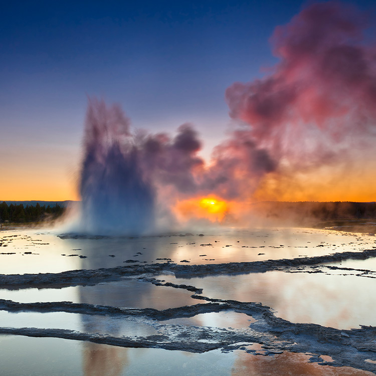 A plume of smoke rising from layers of slate-like liquid at Great Fountain Geyser in Yellowstone National Park at sunset.