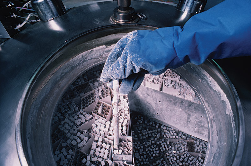 Gloved hand covered in ice, holding frozen vial, above cryopreservation tank filled with boxes of frozen vials.