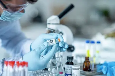 Scientist wearing mask, safety glasses, and gloves looking closely at a small vial in a laboratory setting.