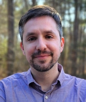 Portrait of Jason Wittenbach wearing a light purple button-down shirt, standing outdoors with trees softly blurred in the background.