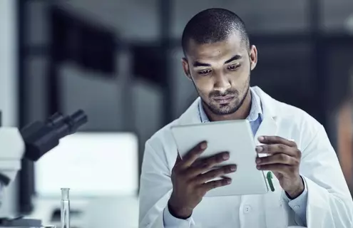 Male scientist in white lab coat, sitting next to microscope, looking at tablet.
