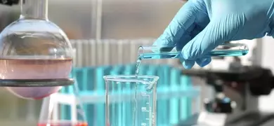 A gloved hand pours blue liquid from a test tube into a graduated beaker in a laboratory, with additional lab equipment and test tubes visible in the background.