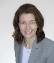 Professional studio portrait of an adult woman wearing a gray blazer over a light-colored top, posed at a slight angle against a neutral light background.