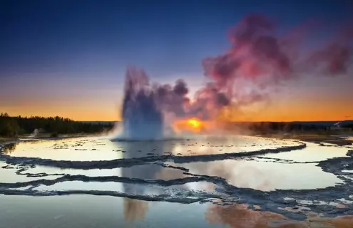 A plume of smoke rising from layers of slate-like liquid at Great Fountain Geyser in Yellowstone National Park at sunset.