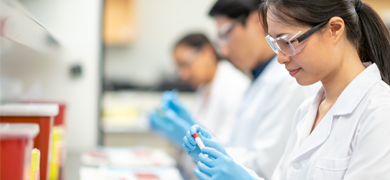 Profile of female scientist wearing safety goggles, lab coat, and gloves looking at sample in test tube, two scientist in background.