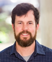 James J Davis wearing a dark blue checkered shirt over a gray undershirt, posing for a professional portrait against a softly blurred neutral background with greenery.