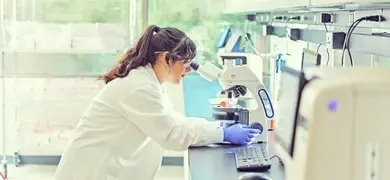 A scientist in a white lab coat and blue gloves looks through a microscope in a laboratory.
