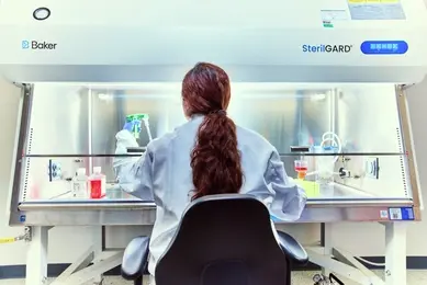 A scientist in a lab coat works under a SterilGARD hood, handling lab equipment and samples on a workbench filled with containers and tools.