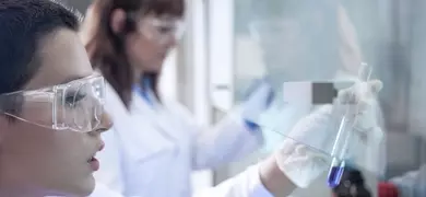 Scientist wearing safety glasses and gloves looking at test tube containing small amount of blue liquid under a hood in a lab.