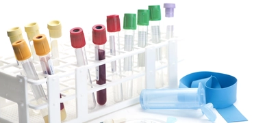 A collection of blood sample tubes in a white plastic rack, each with different colored caps (yellow, red, green, and purple). In front of the rack are medical tools including a blue tourniquet, a needle with tubing, and a clear plastic holder.