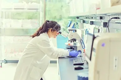 A scientist in a white lab coat and blue gloves looks through a microscope in a laboratory.