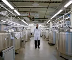 Scientist wearing lab coat, facing camera, standing between rows of large cryopreservation storage tanks at biorepository.