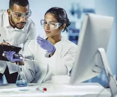Two scientists wearing safety glasses looking at pipette and test tube and sitting in front of computer.