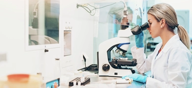 A laboratory professional wearing a white coat and blue gloves is using a microscope at a clean, organized workstation with lab equipment and containers visible on the bench.