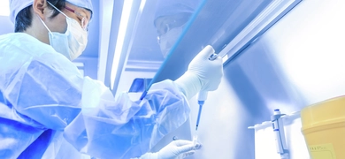 Scientist wearing cap, mask, gown, and gloves holding pipette and vial under a hood.