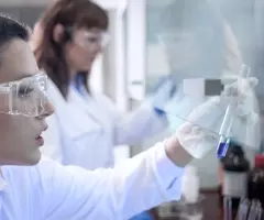 Scientist wearing safety glasses and gloves looking at test tube containing small amount of blue liquid under a hood in a lab.