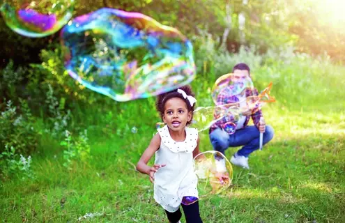 A young Black girl with a ribbon in her hair running outside in the grass toward a huge elongated bubble while a smiling man with a bubble blower squats in the background.