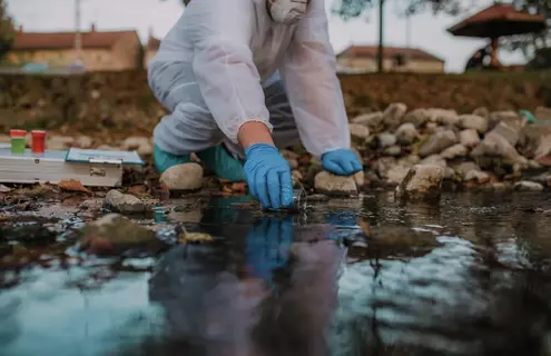 Person in personal protective equipment squatting and reaching to collect a water sample from a stream.