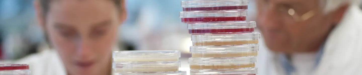 Scientists in lab coats seen over top of stacks of agar plates and small vials.