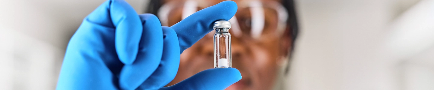 Out of focus black, male, scientist wearing a blue glove holding a Microquant vial infront of their face that is in focus. 