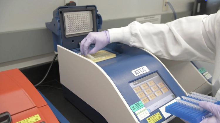 A laboratory technician wearing purple gloves is placing a tray of samples into a PCR machine labeled "ATCC" with a touchscreen interface displaying various options.
