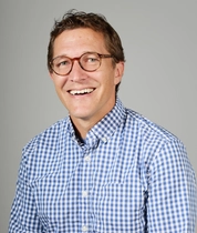 Seated adult man wearing a blue and white checkered button‑down shirt against a neutral gray studio background.
