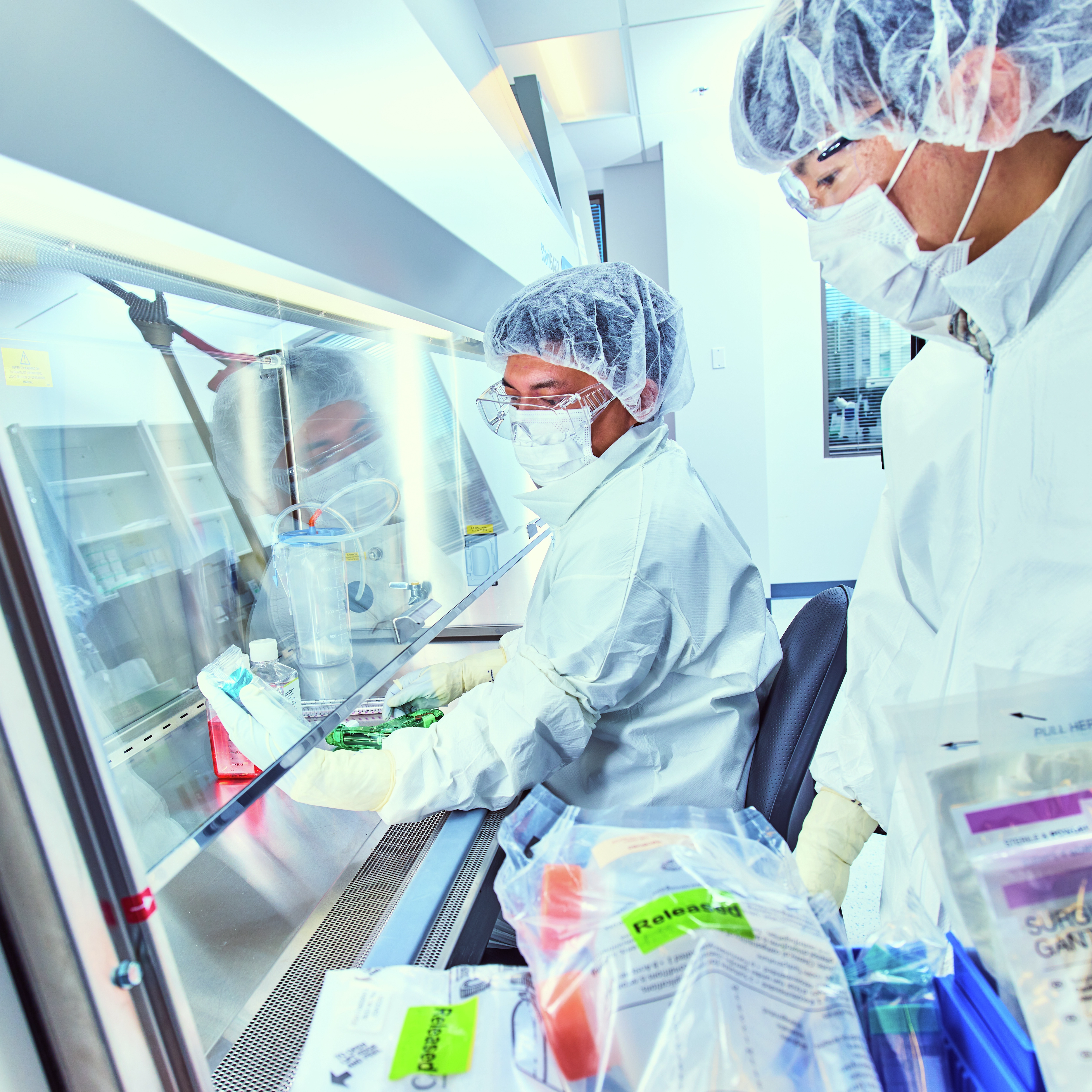 Two ATCC laboratory professionals wearing full cleanroom gowning work at a biosafety cabinet, with one dispensing liquid into a culture container while the other stands nearby observing. Sterile supplies, culture media, and the enclosed stainless‑steel workspace indicate controlled aseptic processing in a laboratory environment.
