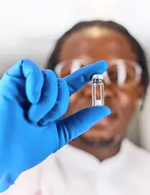 Out of focus black, male, scientist wearing a blue glove holding a Microquant vial infront of their face that is in focus. 